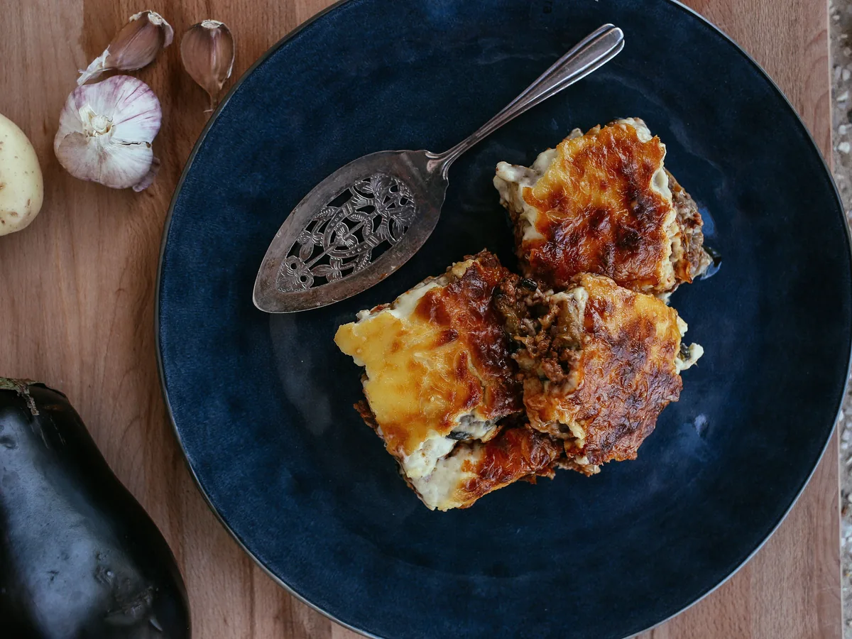 A golden baked eggplant dish with a browned top on a dark blue plate, with an aubergine and garlic beside it